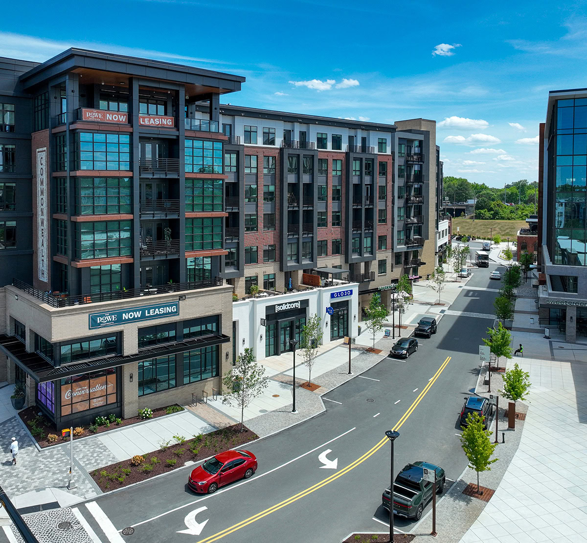 Modern apartment building with balconies and ground-floor retail along a landscaped urban street.