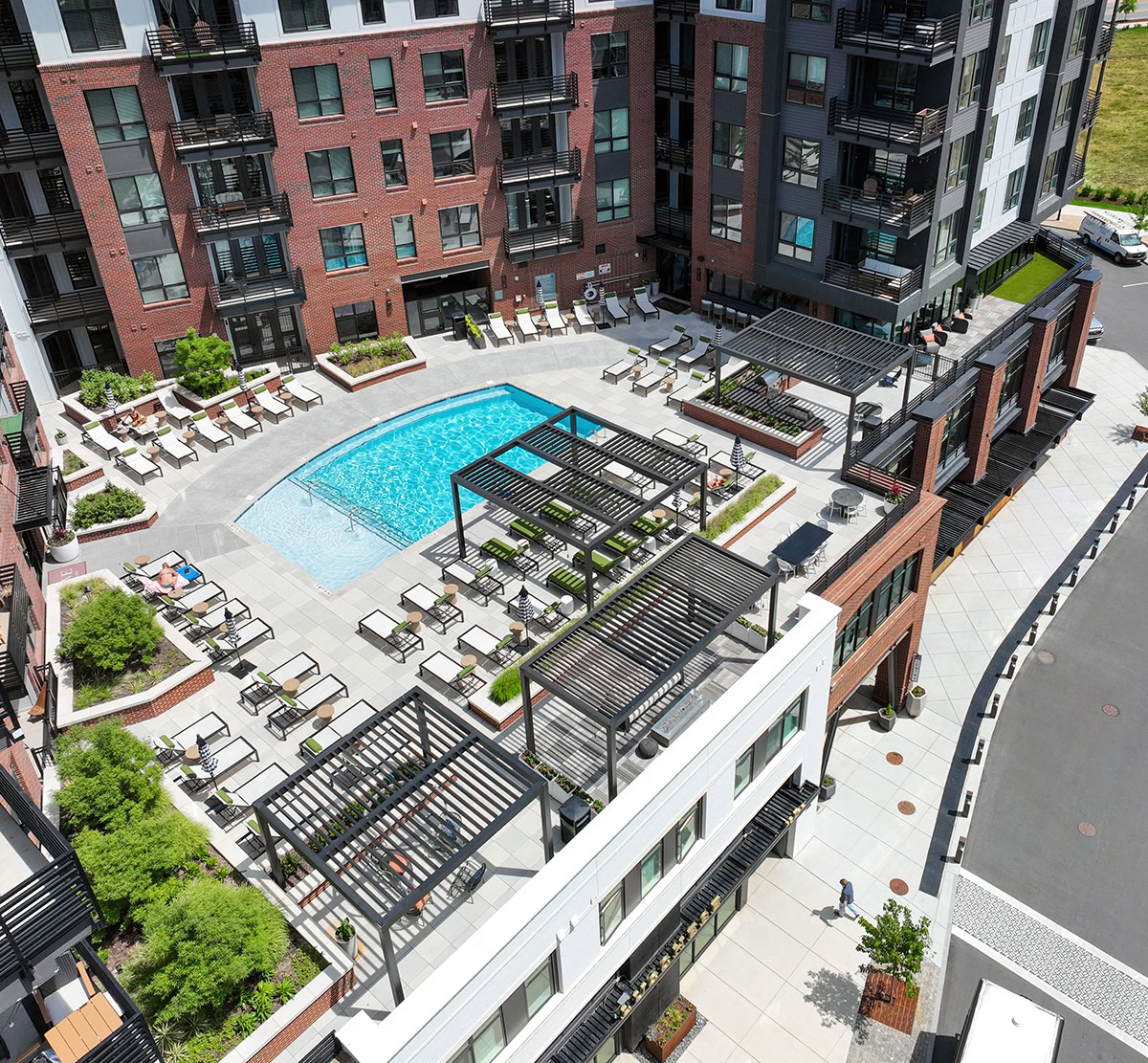 Aerial view of a rooftop pool deck with lounge seating, pergolas, planted areas, and surrounding apartment buildings.