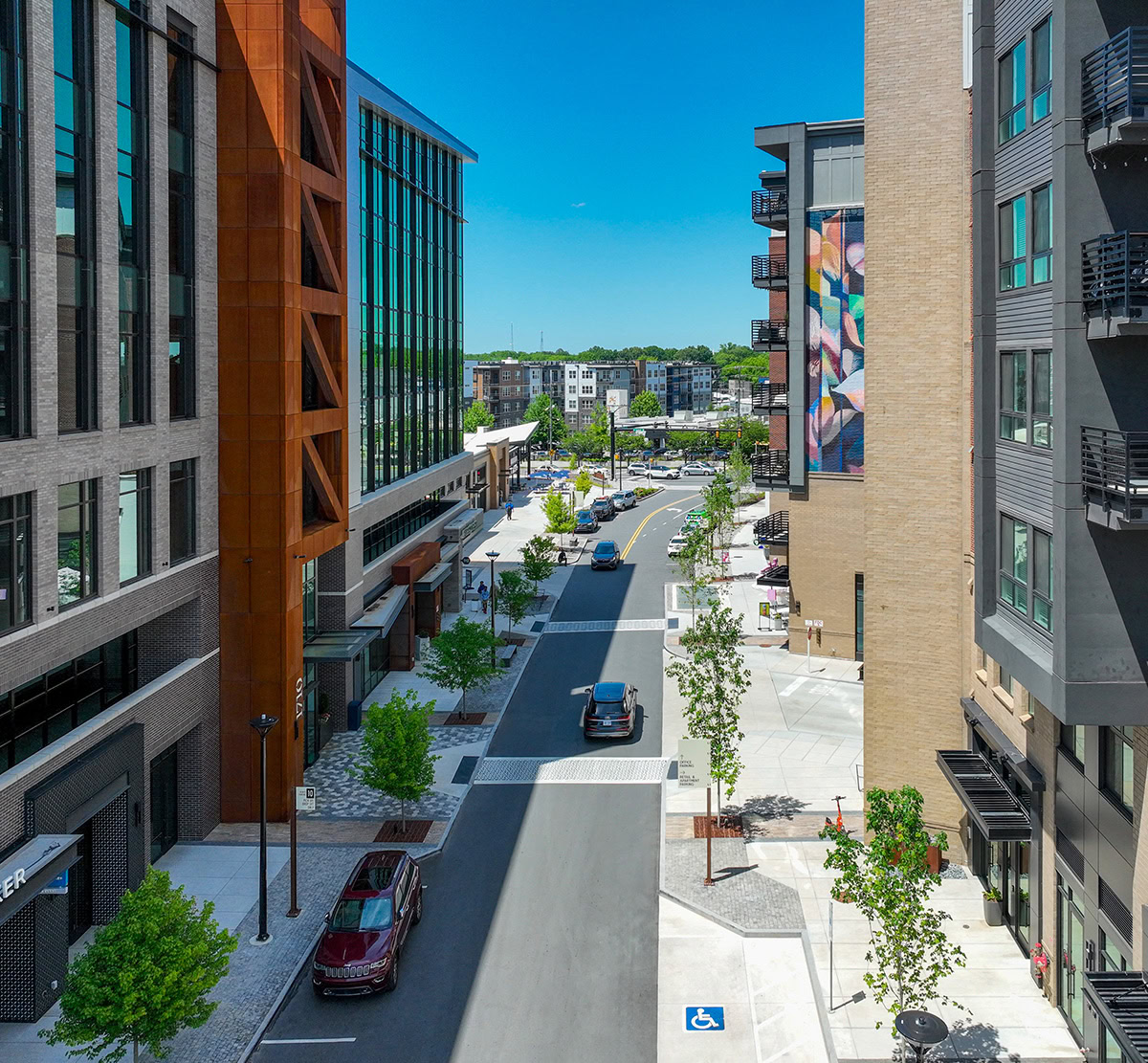 Tree-lined street between modern apartment and office buildings, with sidewalks, storefronts, parked cars, and a colorful wall mural.